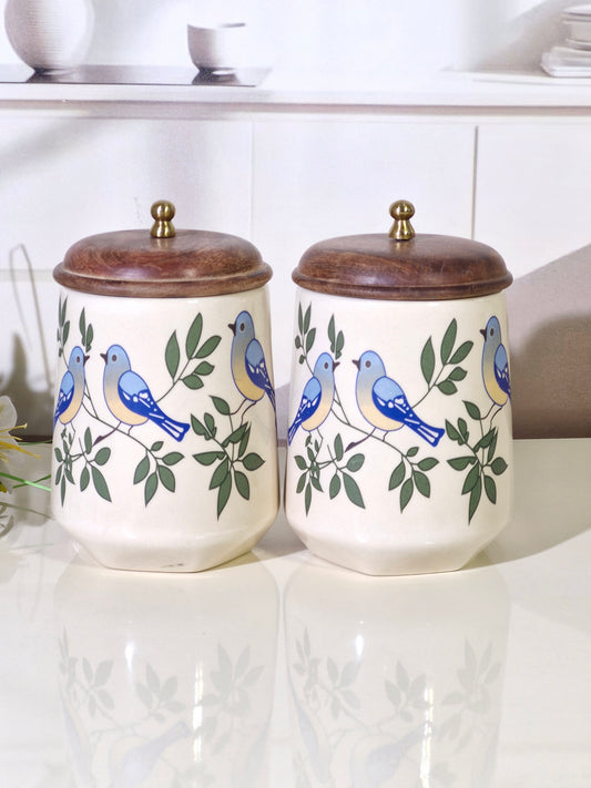 Two cream-colored ceramic canisters with wooden lids, decorated with blue birds and green leaves, displayed on a reflective white surface.