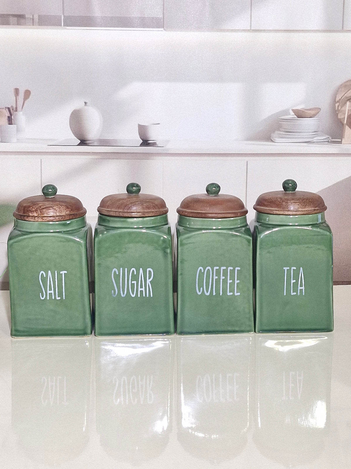 Four green ceramic canisters with copper-colored lids labeled SALT, SUGAR, COFFEE, and TEA, arranged on a glossy white surface in a bright kitchen setting.
