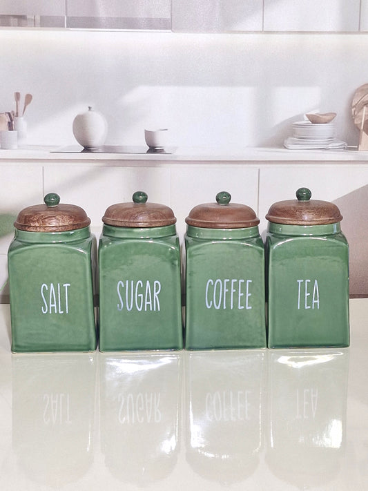 Four green ceramic canisters with copper-colored lids labeled SALT, SUGAR, COFFEE, and TEA, arranged on a glossy white surface in a bright kitchen setting.