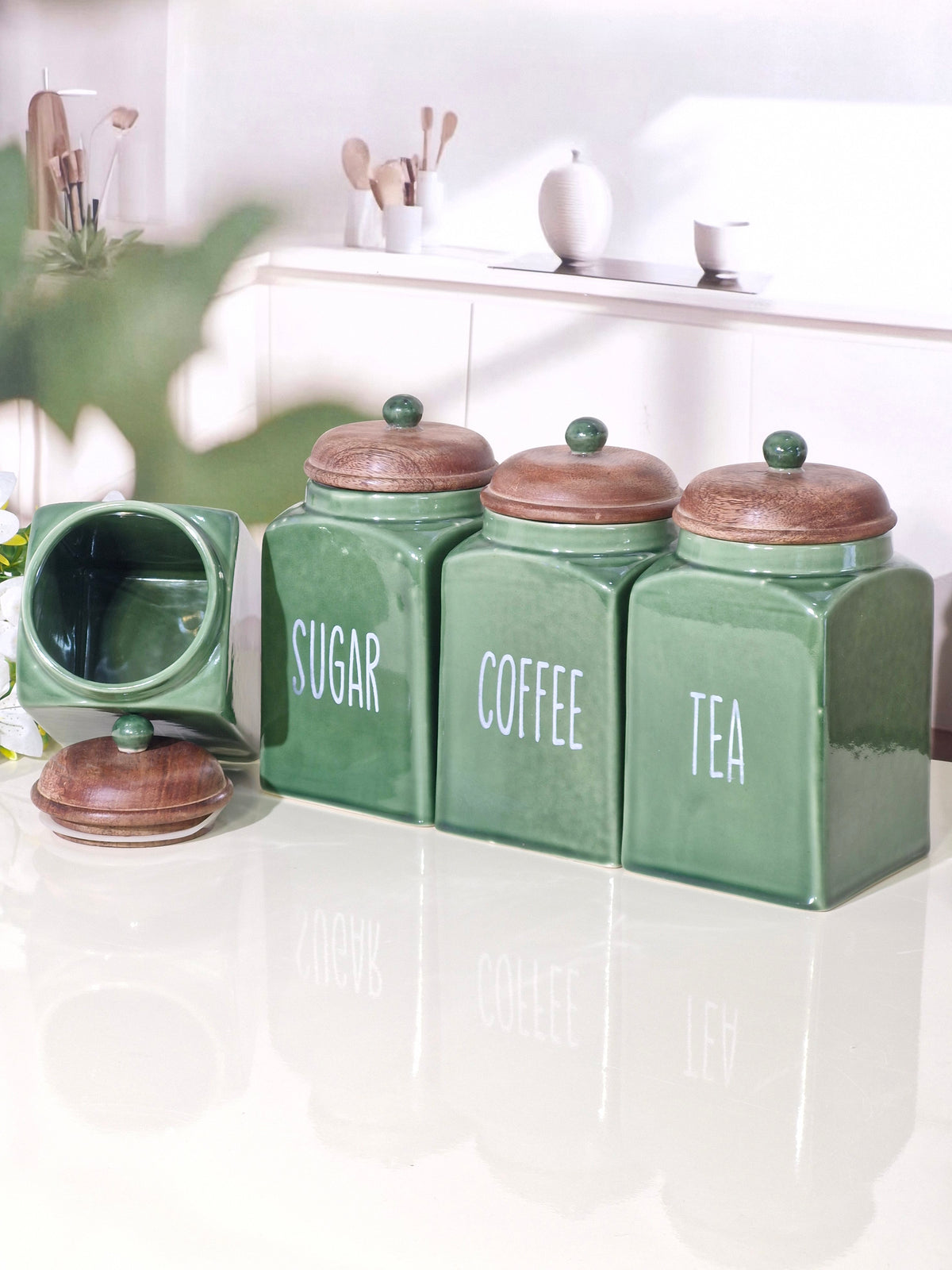 Three green ceramic canisters labeled SUGAR, COFFEE, and TEA with brown wooden lids, alongside a fourth canister with its lid off, set on a glossy white countertop in a bright kitchen.