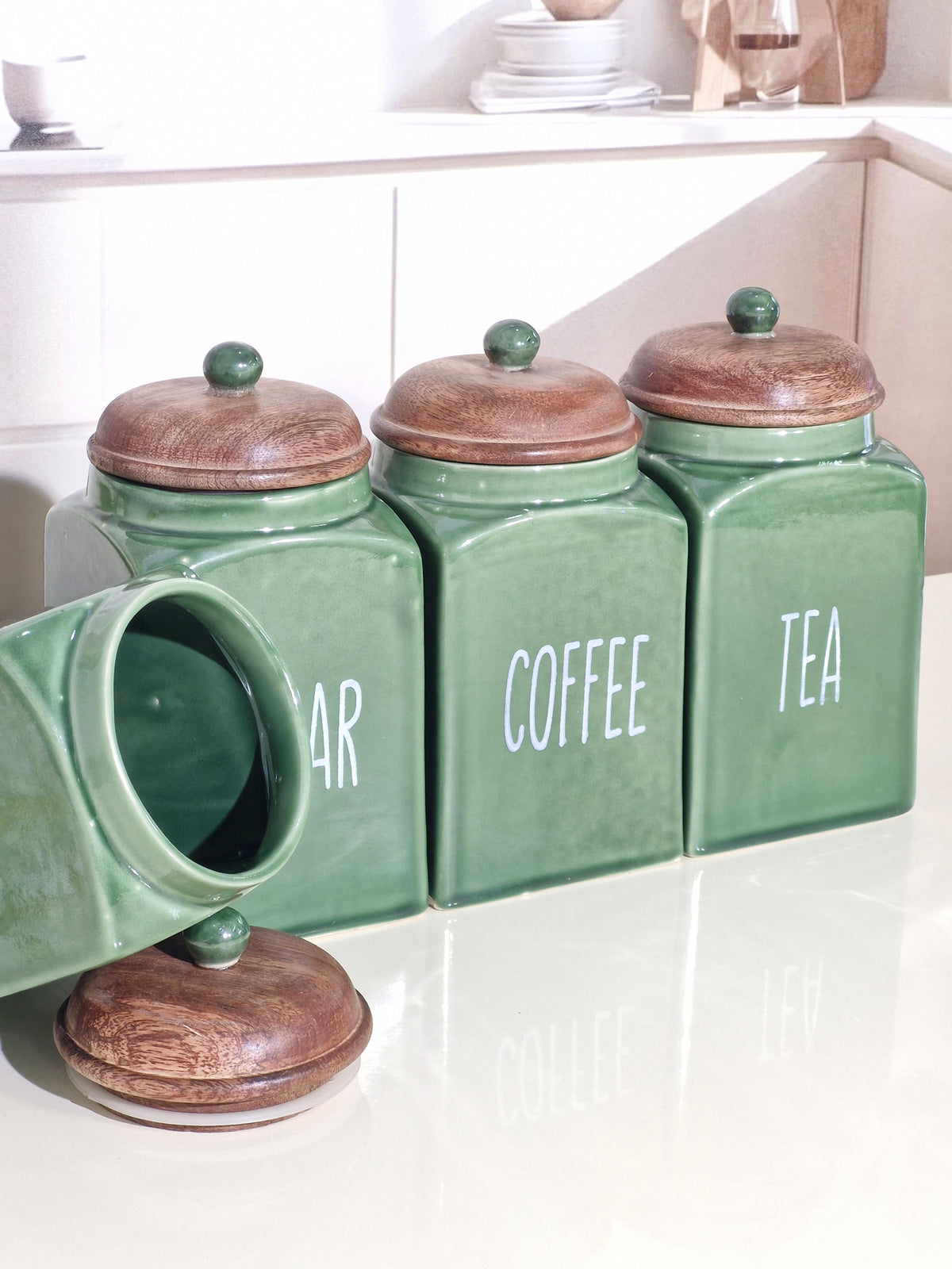 Three green ceramic canisters with wooden lids labeled Sugar, Coffee, and Tea, arranged on a white countertop in a bright kitchen setting.