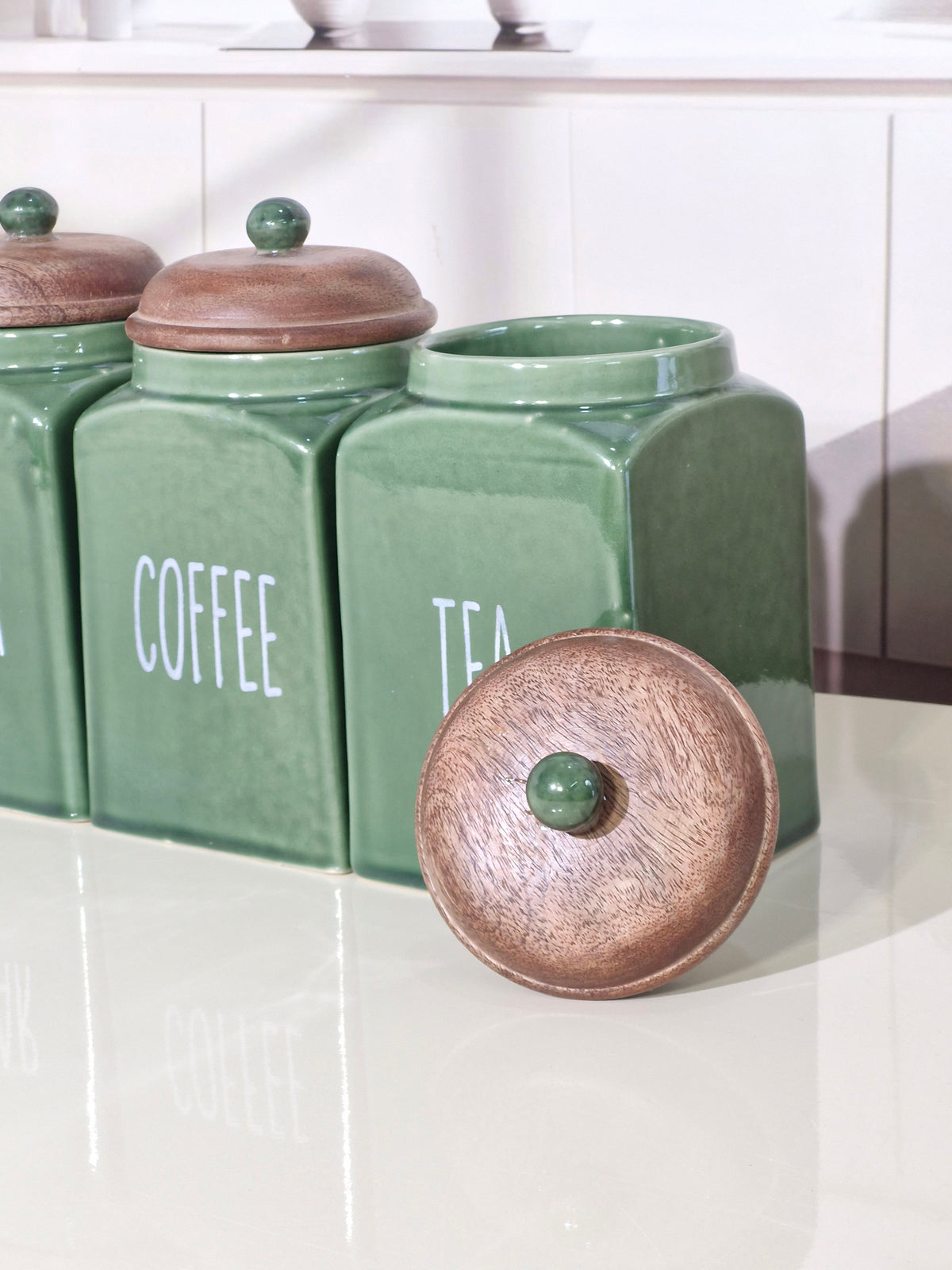 Three green ceramic canisters labeled COFFEE, TEA, and another canister, with one copper-toned lid placed in front on a glossy white surface.