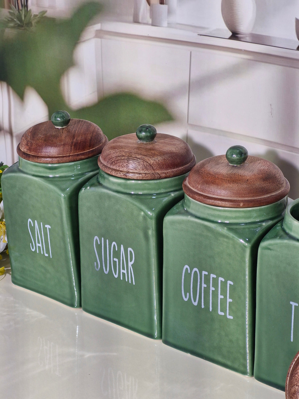 Four green ceramic canisters with wooden lids and white lettering for Salt, Sugar, Coffee, and another canister, arranged on a countertop in a kitchen setting.