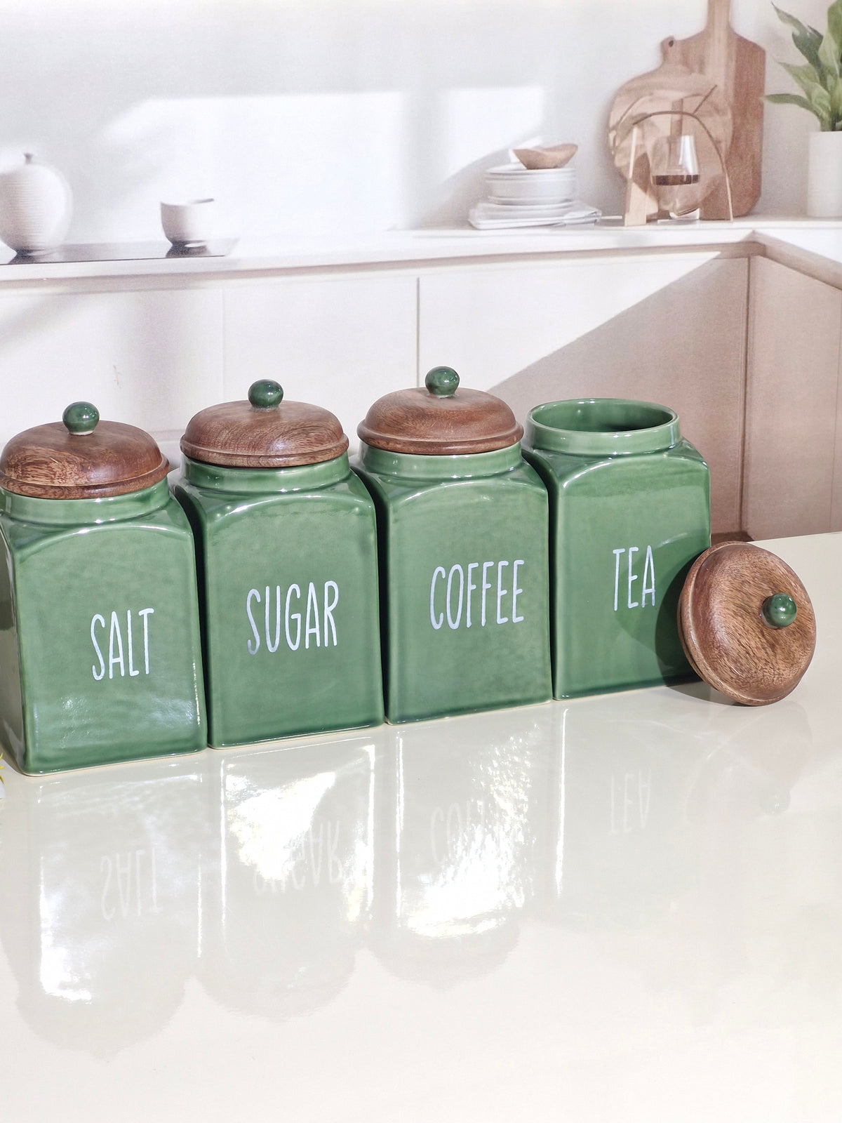 Four green ceramic canisters with wooden lids labeled Salt, Sugar, Coffee, and Tea, arranged on a glossy white countertop in a bright kitchen setting.