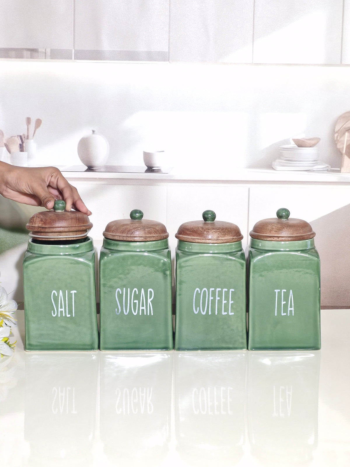 Four green ceramic canisters labeled SALT, SUGAR, COFFEE, and TEA with brown wooden lids, arranged on a white countertop in a bright kitchen setting. A hand is touching the leftmost canister.