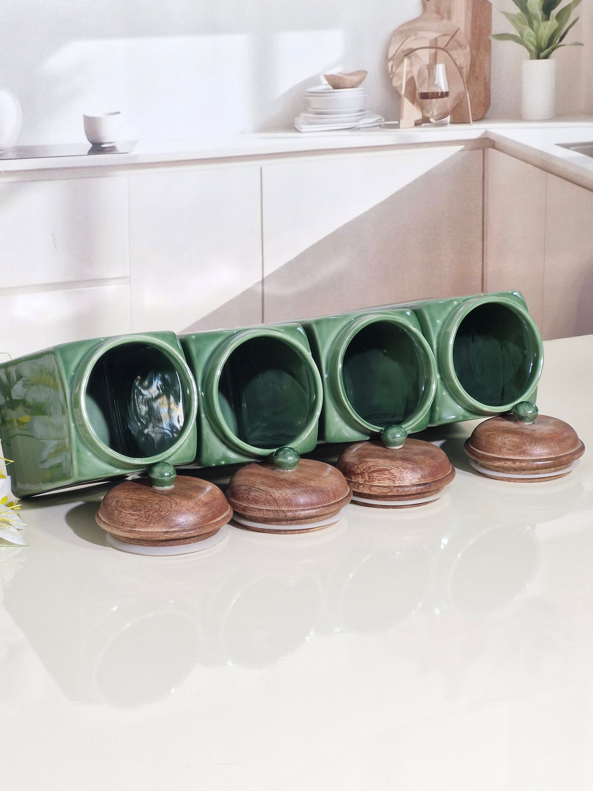 Four green ceramic spice jars with matching wooden lids arranged on a glossy white countertop in a bright kitchen setting.