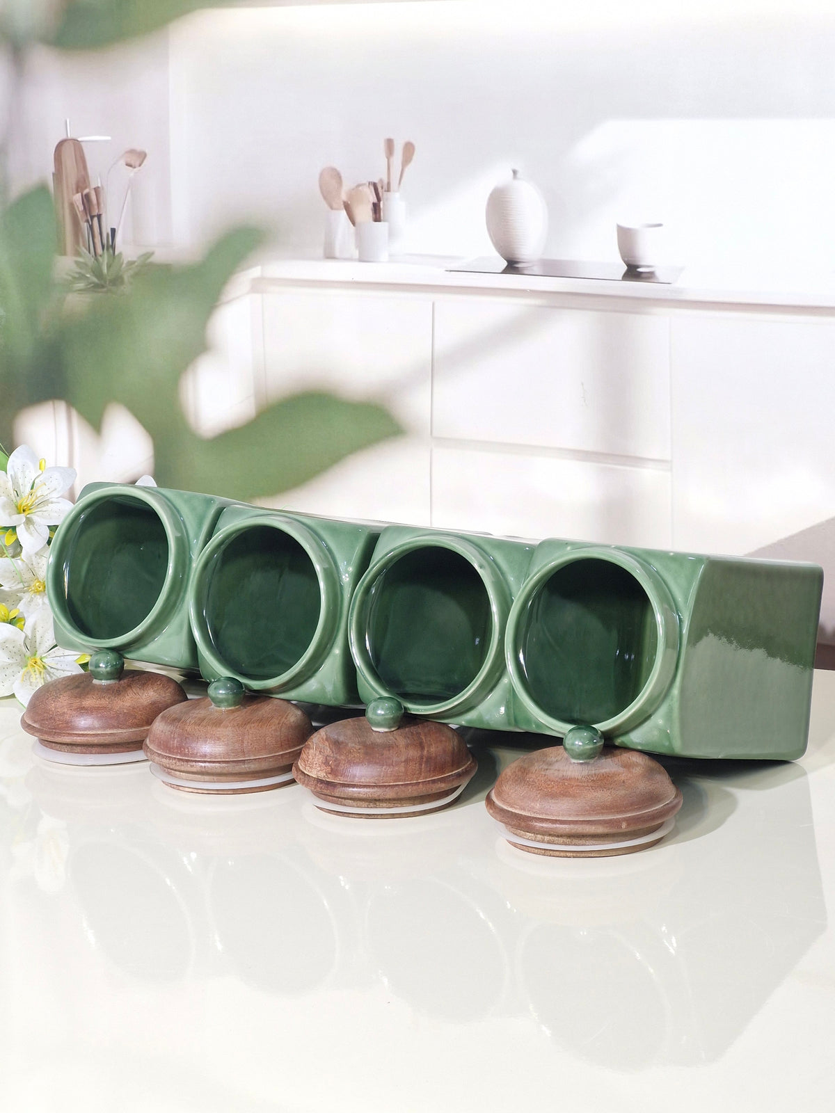 A green ceramic spice rack with four round hanging hooks and four matching lidded jars, displayed on a glossy white surface in a bright kitchen setting.