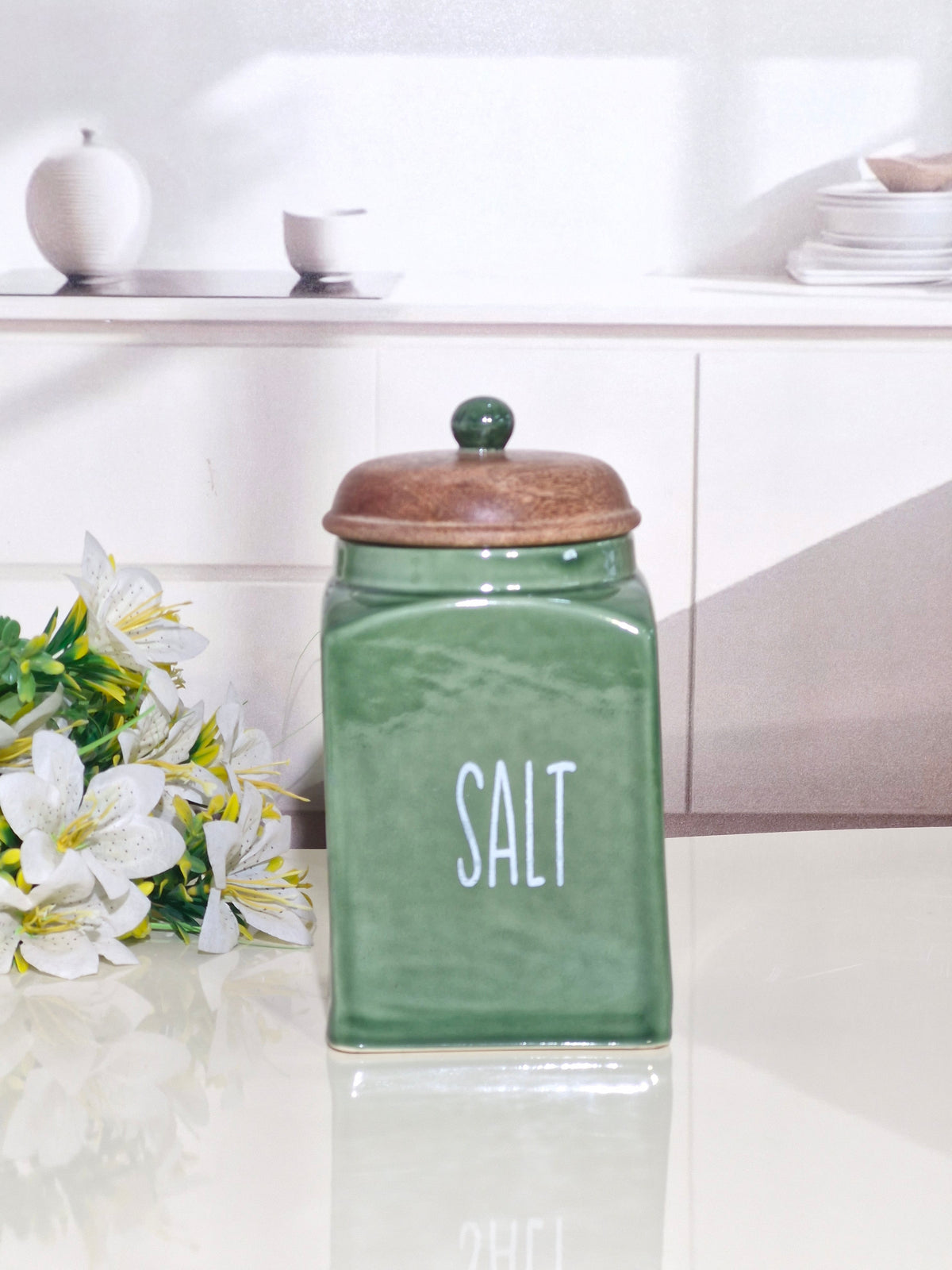 A green ceramic salt canister with a wooden lid and white 'SALT' text, set on a white countertop with white flowers and a blurred kitchen background.