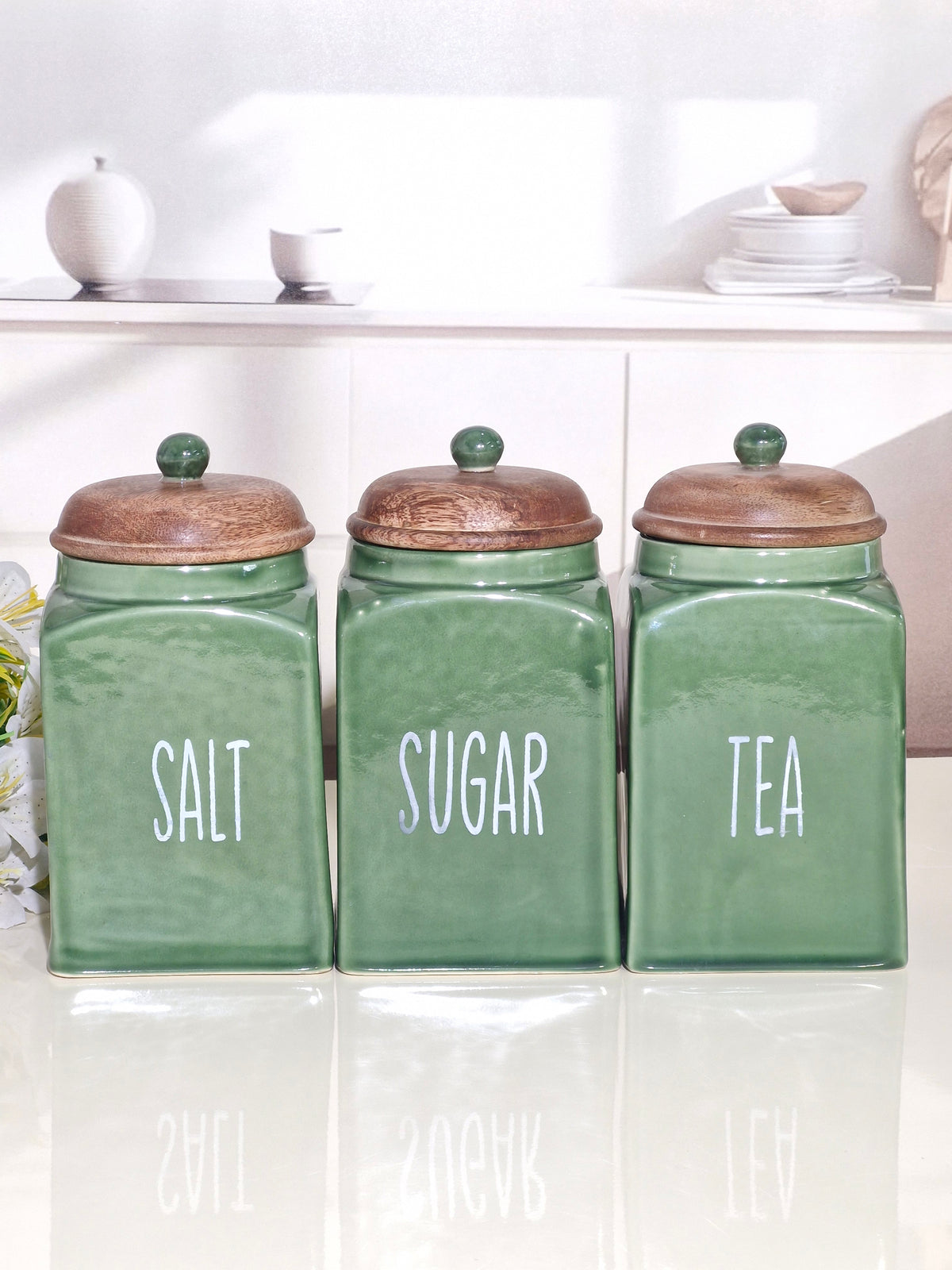 Three green ceramic canisters with wooden lids labeled SALT, SUGAR, and TEA, arranged on a glossy white surface in a bright kitchen setting.