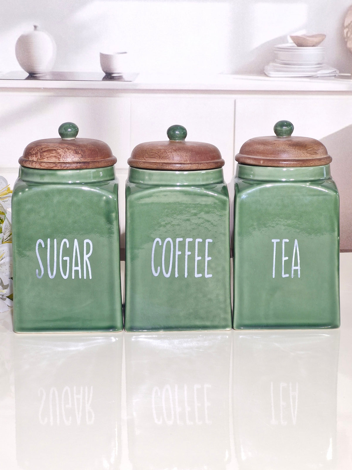 Three green ceramic canisters with wooden lids labeled SUGAR, COFFEE, and TEA, arranged on a glossy white surface in a bright kitchen setting.