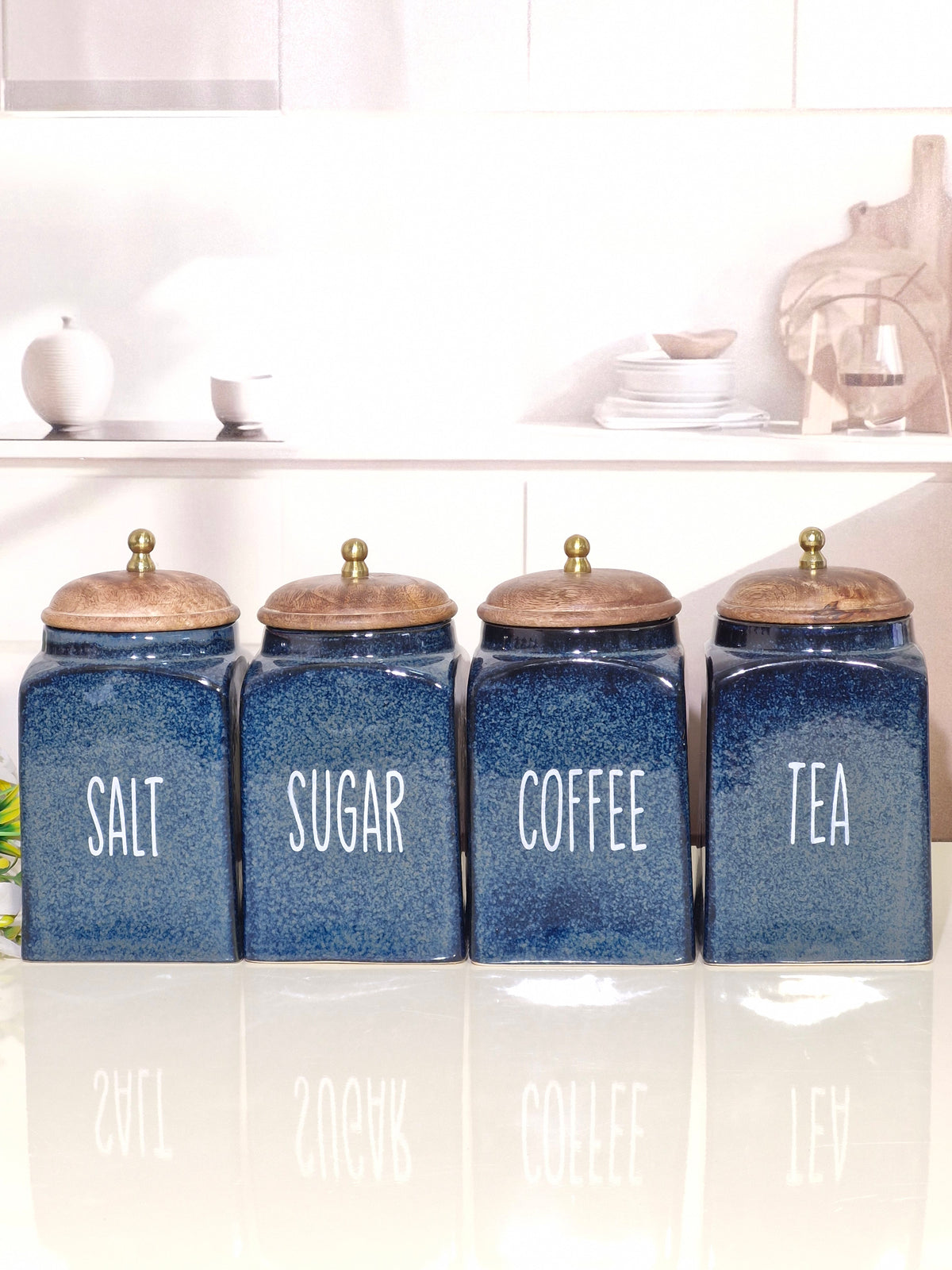 Four blue ceramic canisters with wooden lids and white lettering for Salt, Sugar, Coffee, and Tea, lined up on a reflective white surface in a bright kitchen setting.