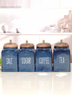 Four blue ceramic canisters with wooden lids and white lettering for Salt, Sugar, Coffee, and Tea, lined up on a reflective white surface in a bright kitchen setting.