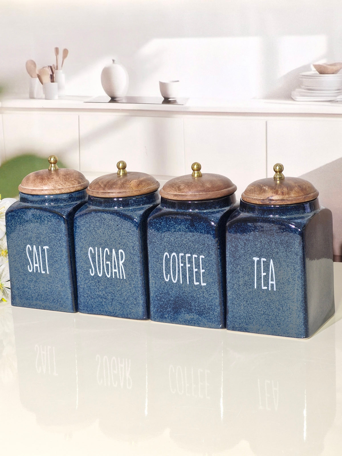 Four blue ceramic canisters with wooden lids and brass knobs, labeled Salt, Sugar, Coffee, and Tea, lined up on a glossy white surface.
