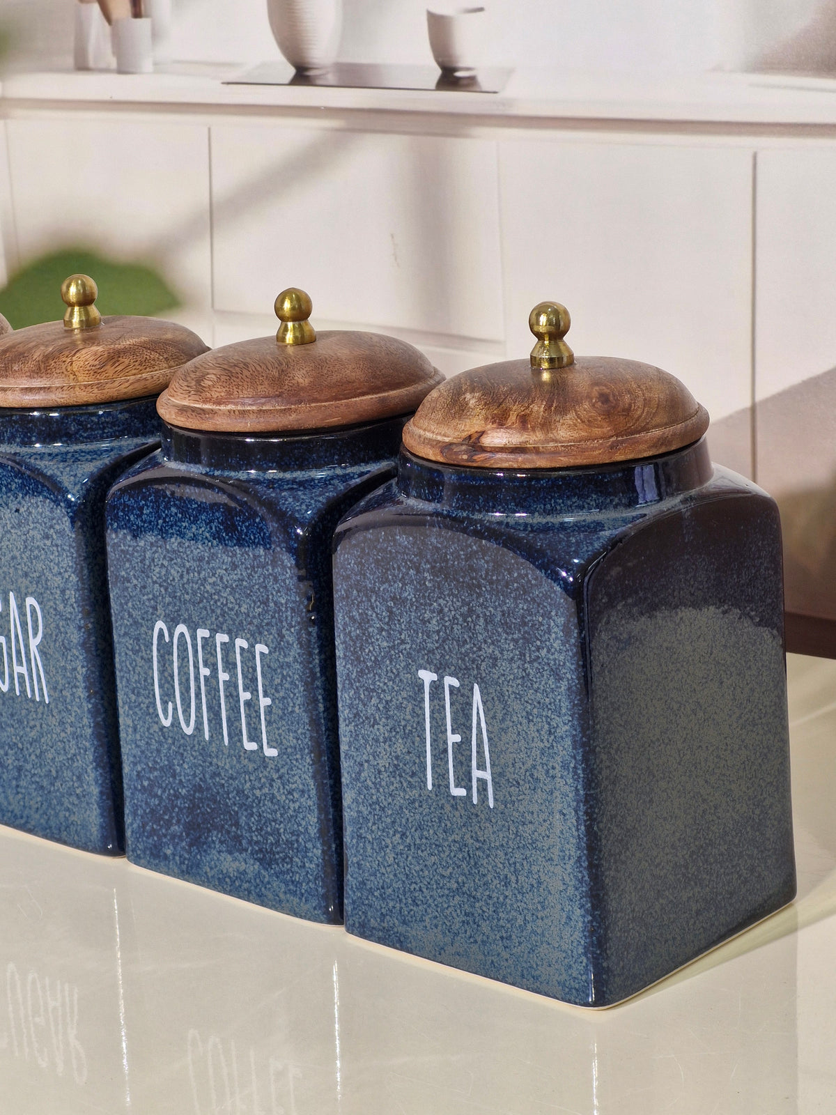 Three blue ceramic tea and coffee canisters with wooden lids and brass knobs, labeled 'SUGAR', 'COFFEE', and 'TEA' on a kitchen counter.