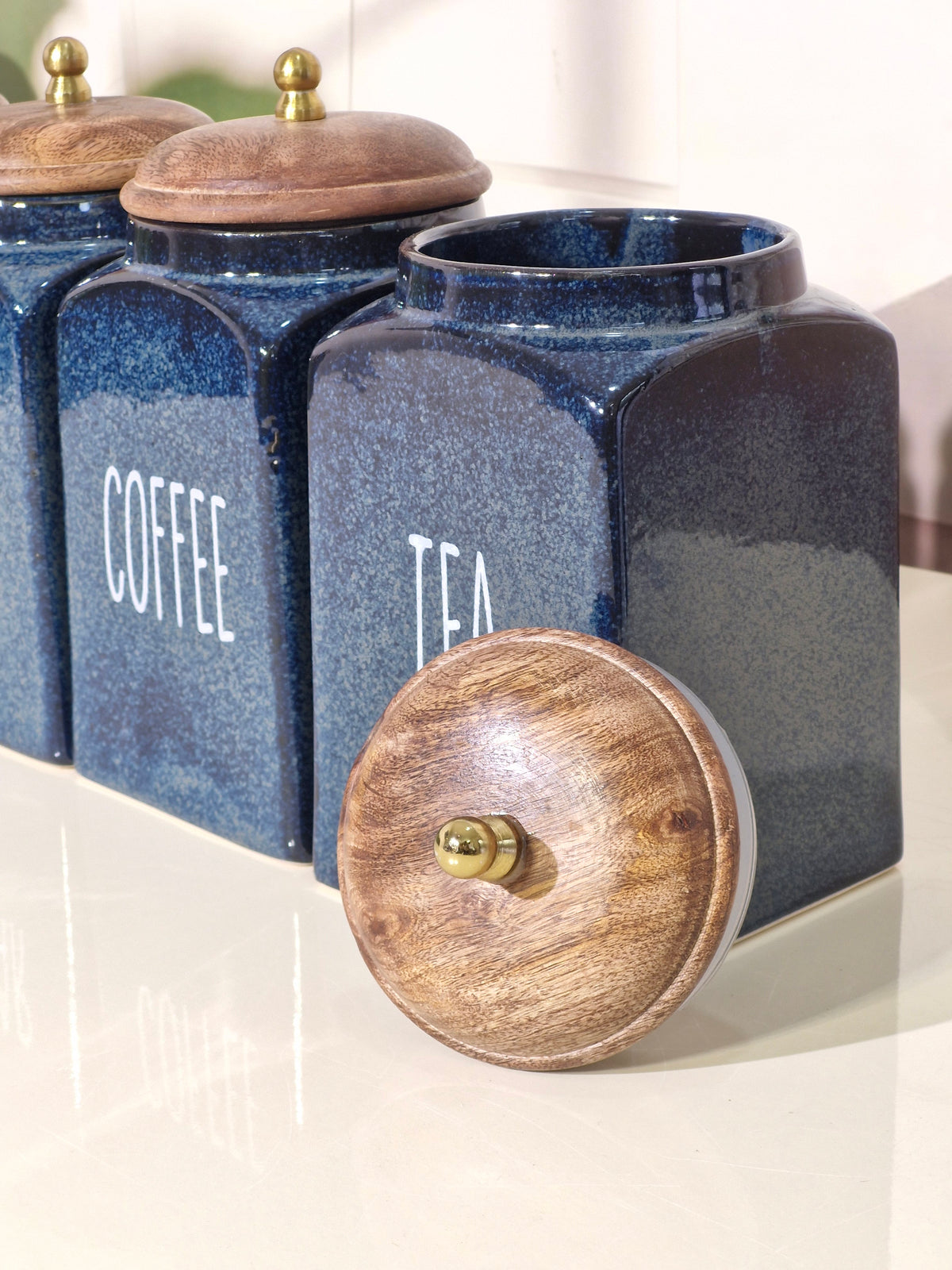 Three blue ceramic canisters with wooden lids on a glossy white surface. The canisters are labeled COFFEE, TEA, and an empty canister in the foreground, with one wooden lid leaning in front.