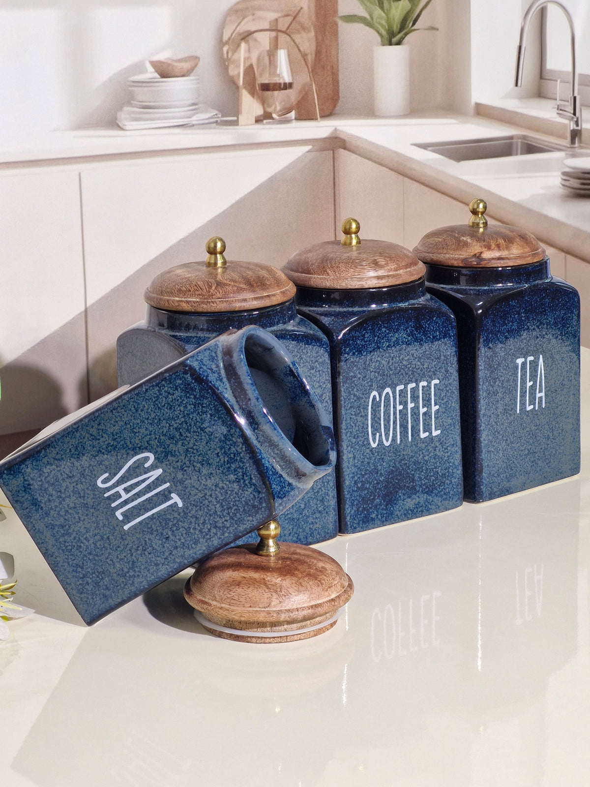 Four blue ceramic canisters with wooden lids and white lettering for Salt, Coffee, Tea, and another canister, arranged on a glossy kitchen countertop.