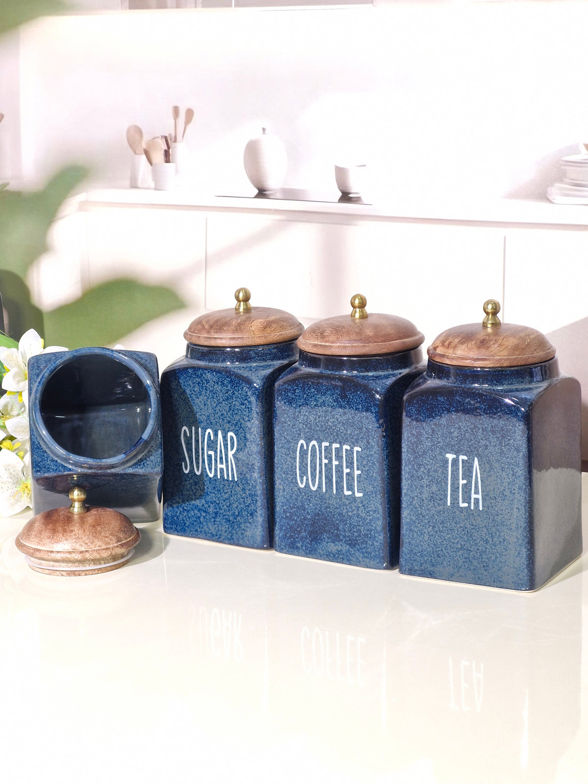 Four blue ceramic canisters labeled SUGAR, COFFEE, and TEA with wooden lids, plus an empty matching canister and a small saucer, arranged on a white surface in a bright kitchen setting.
