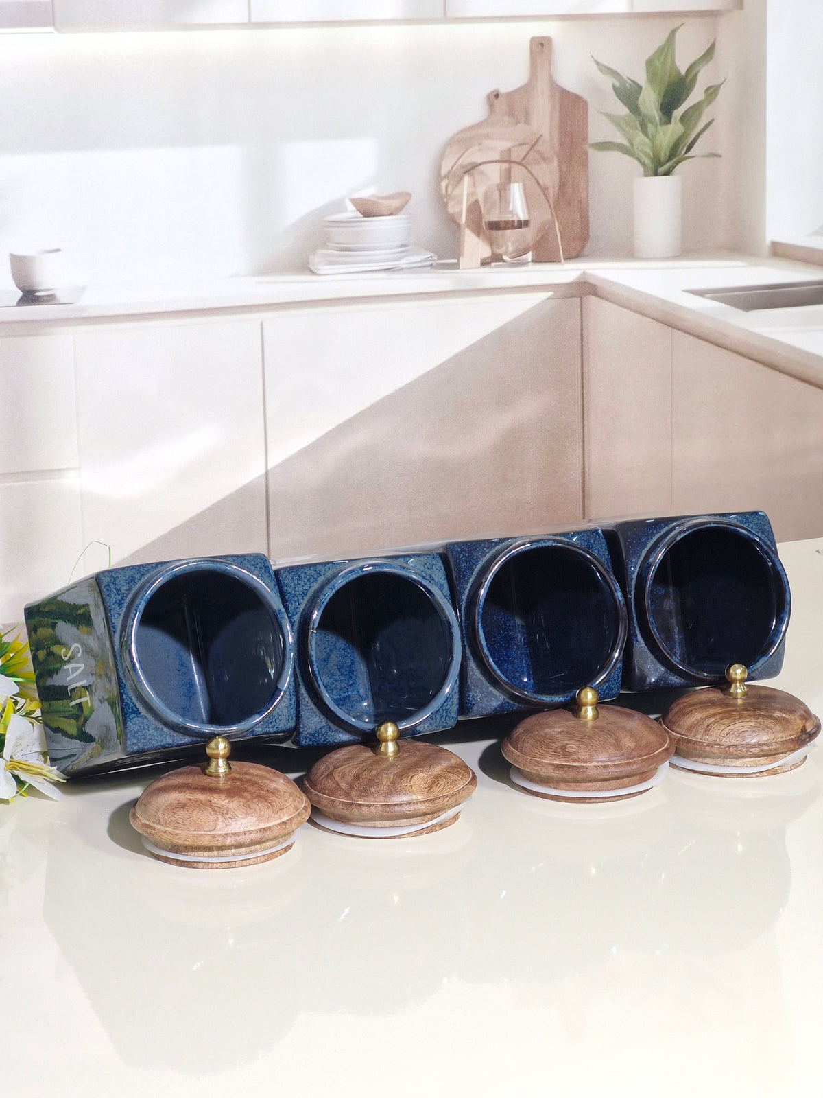 four dark blue ceramic spice jars with lids, arranged in a row on a light countertop in a bright kitchen setting.