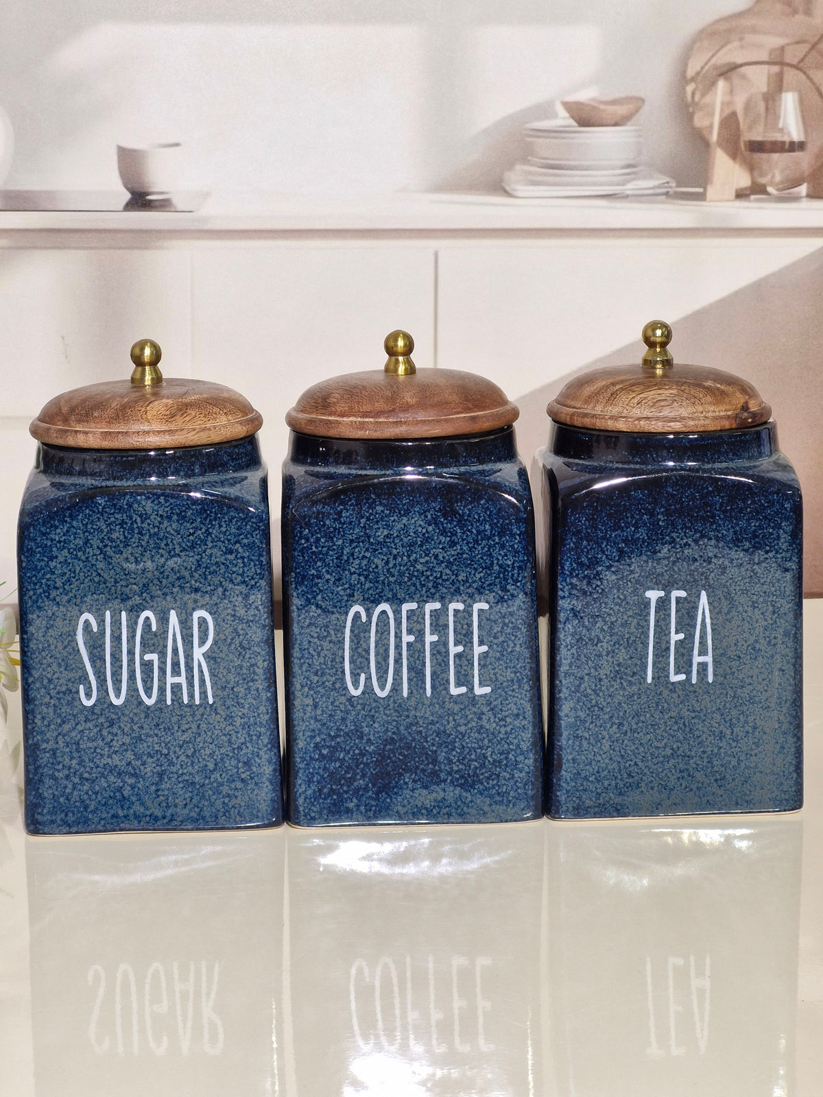 Three blue ceramic canisters labeled SUGAR, COFFEE, and TEA with wooden lids and brass knobs, arranged on a reflective white surface.
