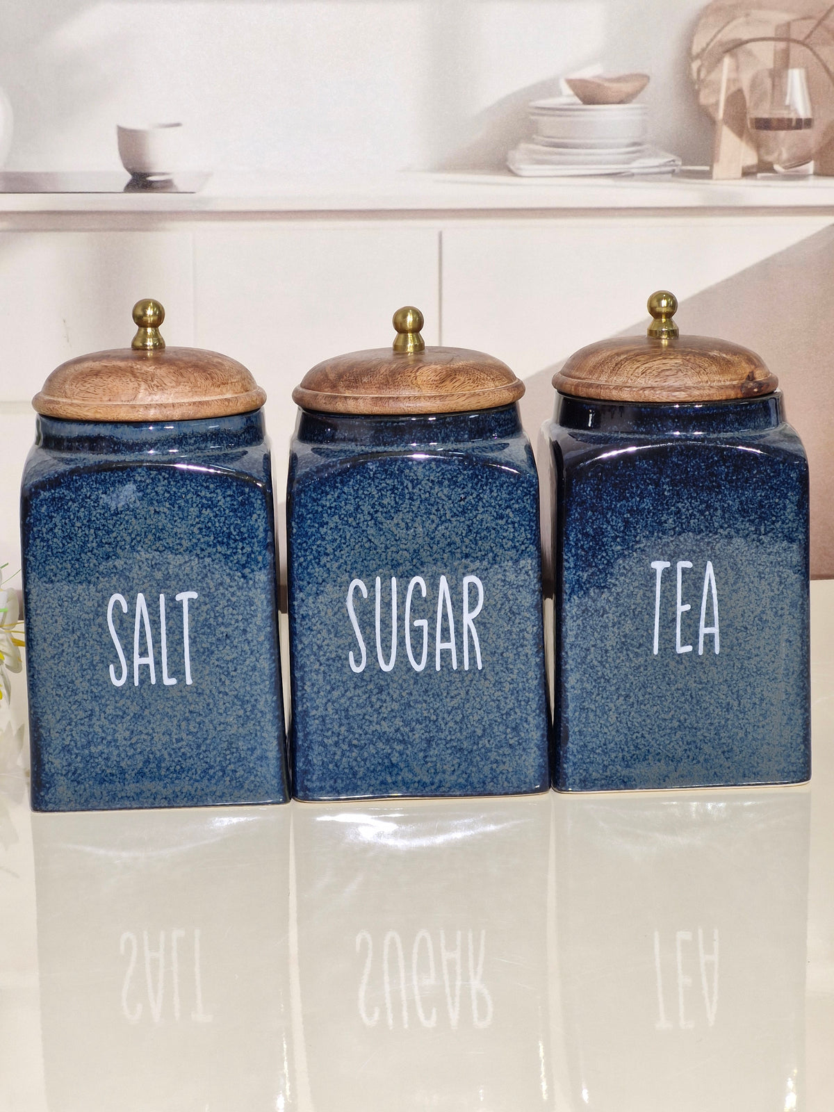 Three blue speckled ceramic canisters labeled SALT, SUGAR, and TEA with wooden lids, arranged on a glossy white surface in a kitchen setting.