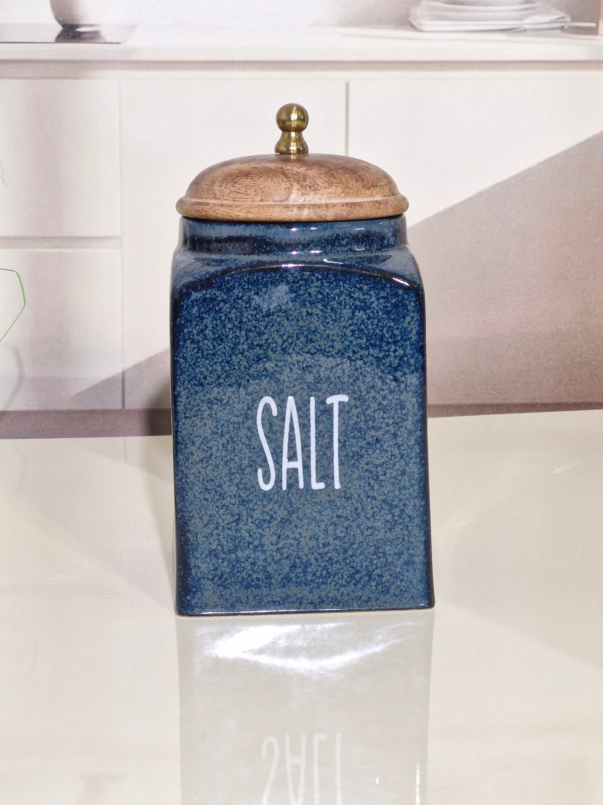 A blue speckled ceramic salt canister with a wooden lid and brass knob, standing on a reflective white surface in a kitchen setting.