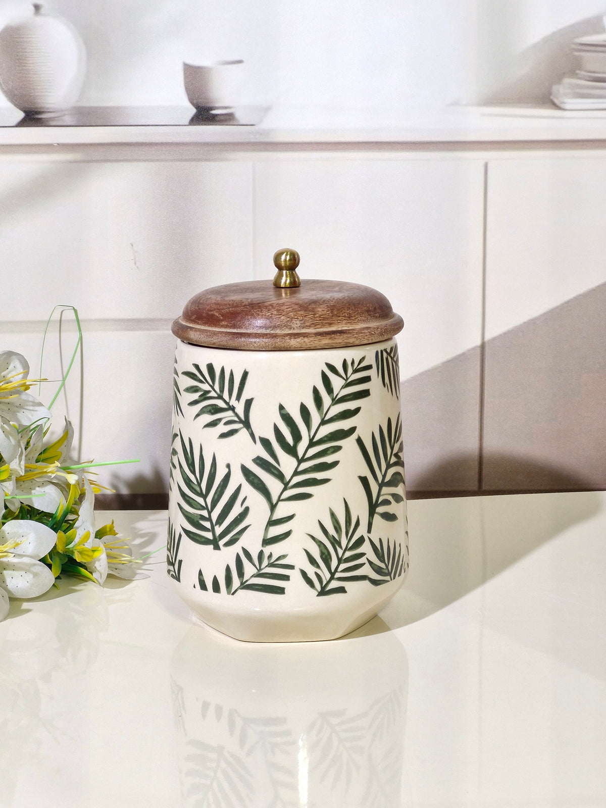 A ceramic canister with a lid, featuring a cream-colored body decorated with a dark green palm leaf pattern and a rustic brown wooden lid with a small brass knob, set on a glossy white countertop.