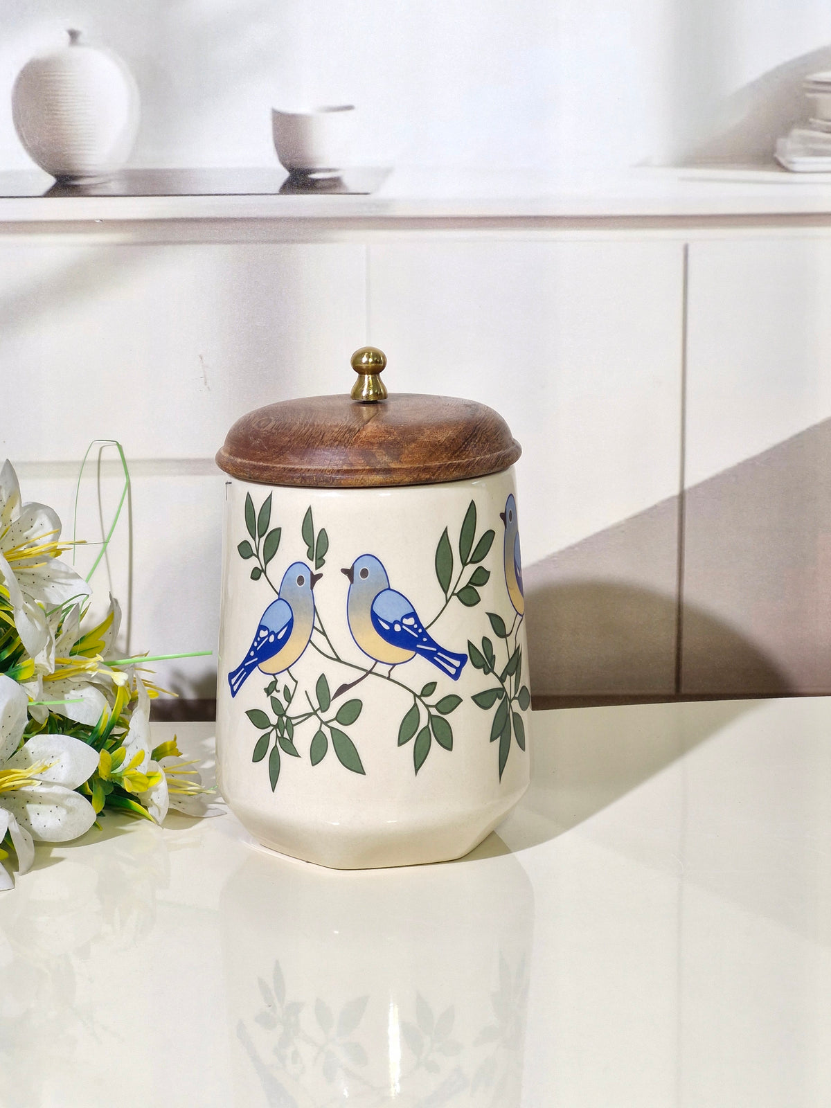 A ceramic canister with a wooden lid, decorated with blue birds and green leaves on a cream background, set on a white countertop with a blurred kitchen background.
