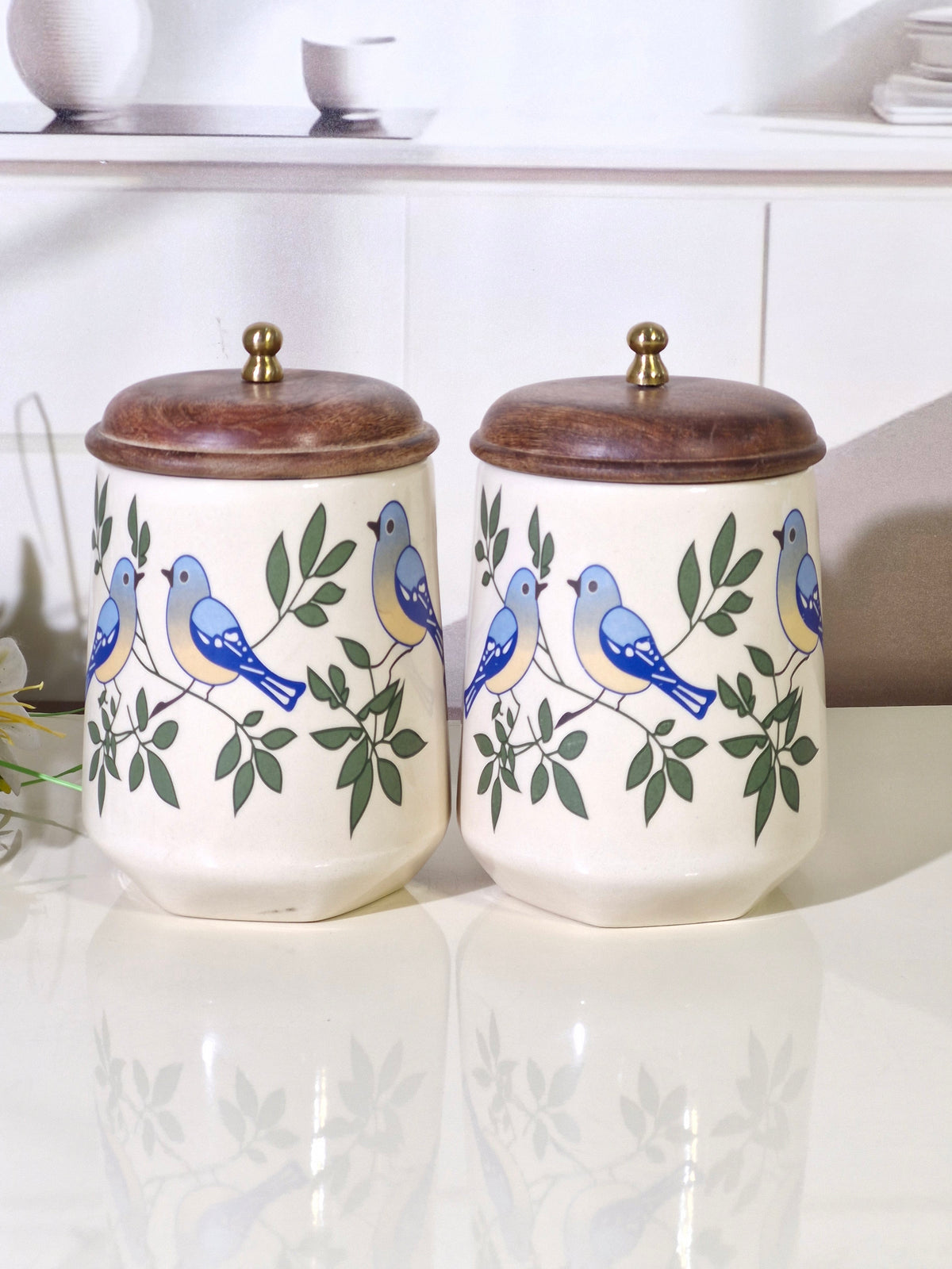 Two cream-colored ceramic canisters with wooden lids, decorated with blue birds and green leaves, displayed on a reflective white surface.