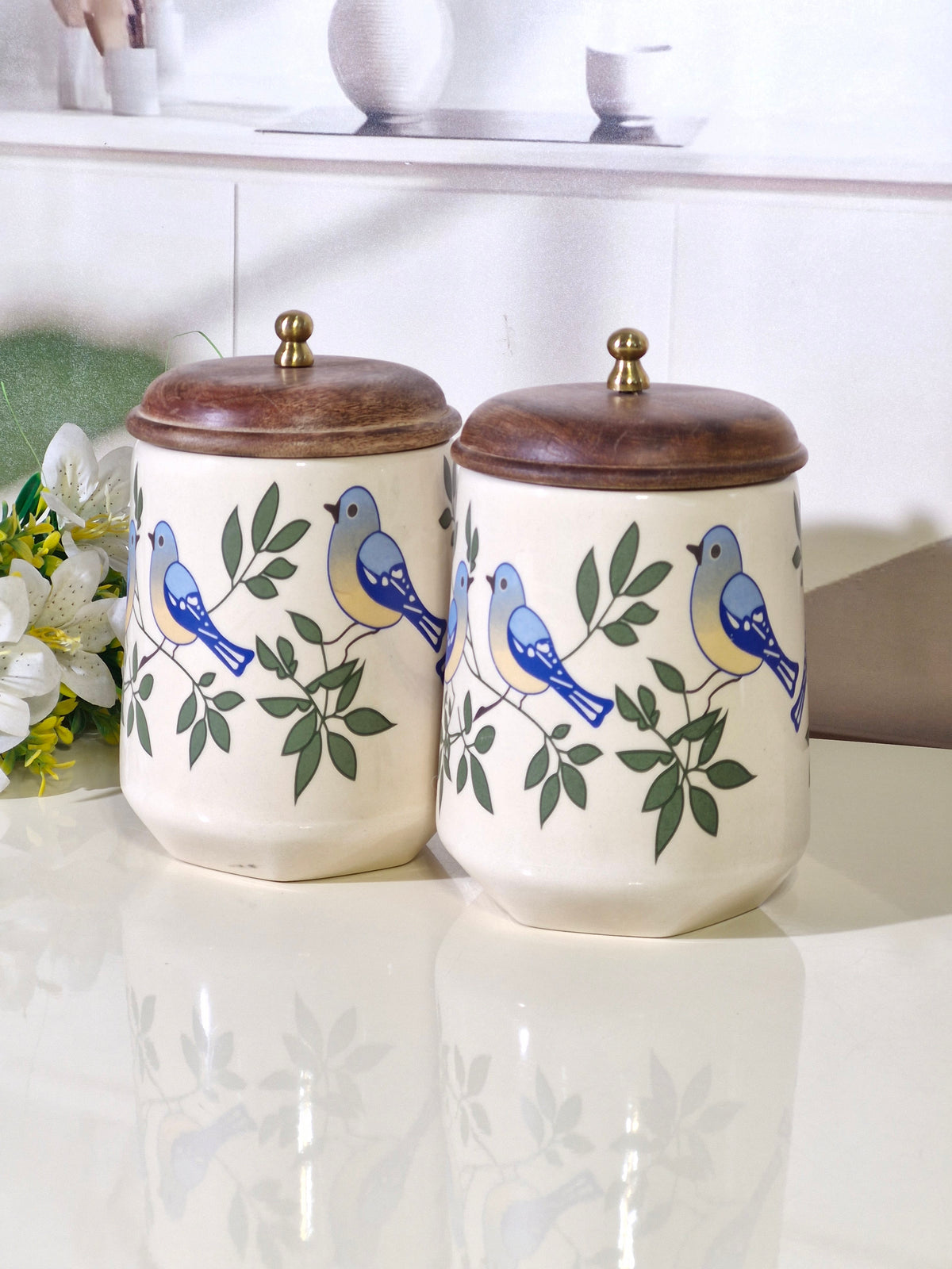 Two cream-colored ceramic canisters with brown wooden lids and brass knobs, decorated with blue birds and green leaves, set on a glossy white surface.