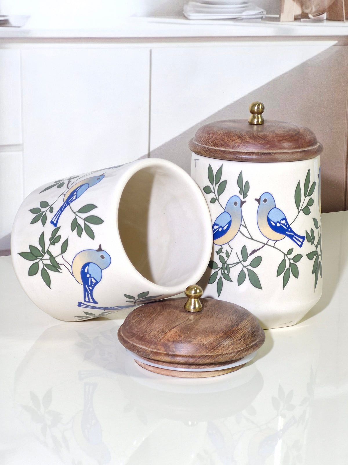 Two ceramic canisters with wooden lids and blue bird illustrations, displayed on a glossy white surface with a kitchen background.