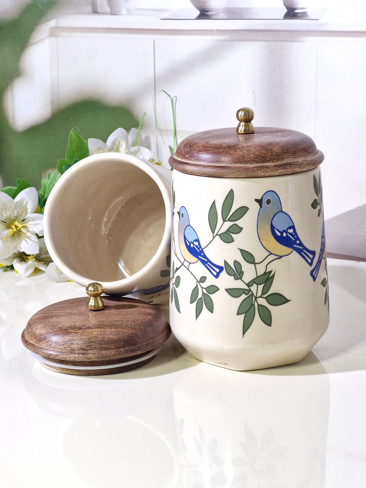 A ceramic canister with a lid, shown closed and open on a glossy white surface. The canister has a cream-colored body decorated with blue birds and green leaves, topped with a dark brown wooden lid and a small brass knob.