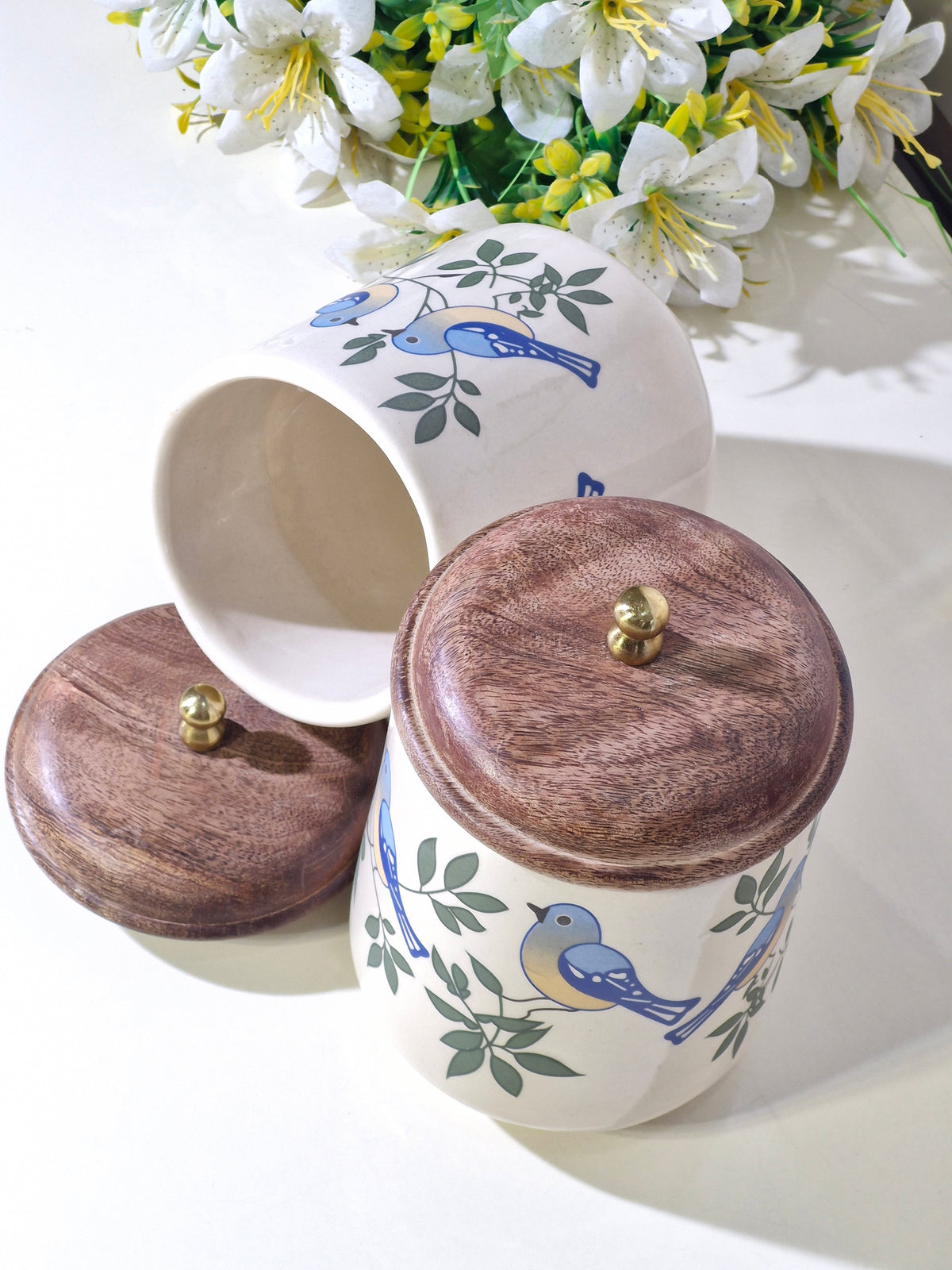 A ceramic canister with a wooden lid and blue bird and green leaf illustrations, shown with its lid off and another canister in the background. White flowers are visible in the upper background.