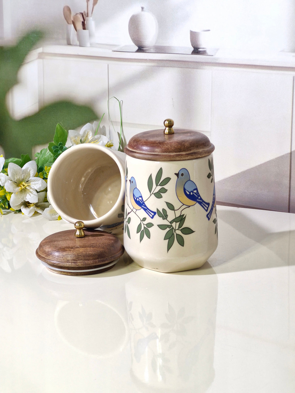 A ceramic canister with a matching lid, decorated with blue birds and green leaves, shown next to an open canister on a glossy white surface.