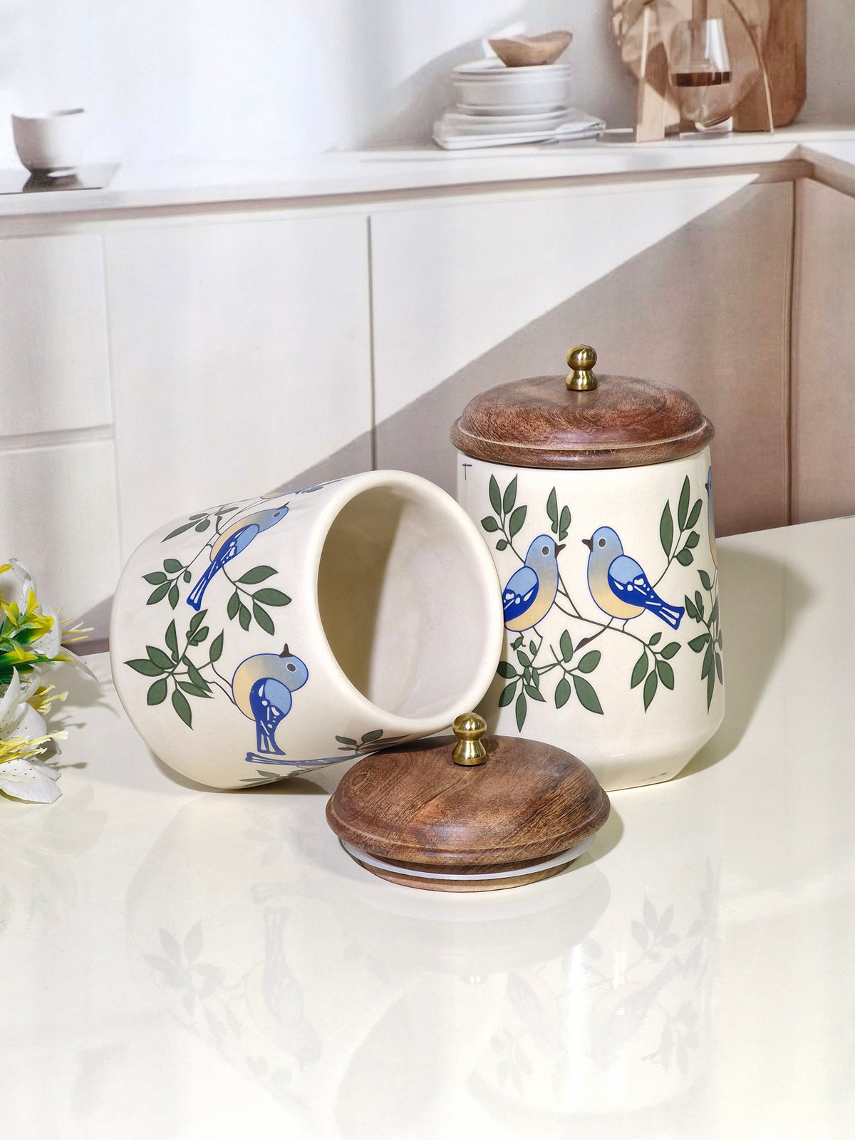 Two ceramic canisters with blue birds and green leaves on a glossy white surface, featuring dark brown wooden lids with brass knobs. One canister is standing upright and the other is lying on its side to show the open interior.