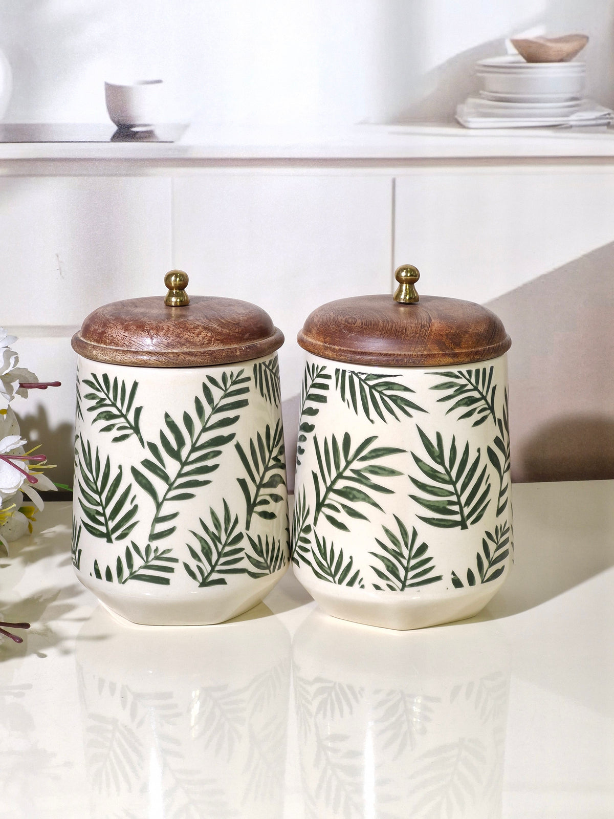 Two ceramic canisters with a green palm leaf pattern on an off-white background, topped with rustic brown wooden lids and small brass knobs, displayed on a glossy white surface.