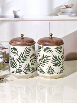 Two ceramic canisters with a green palm leaf pattern on an off-white background, topped with rustic brown wooden lids and small brass knobs, displayed on a glossy white surface.