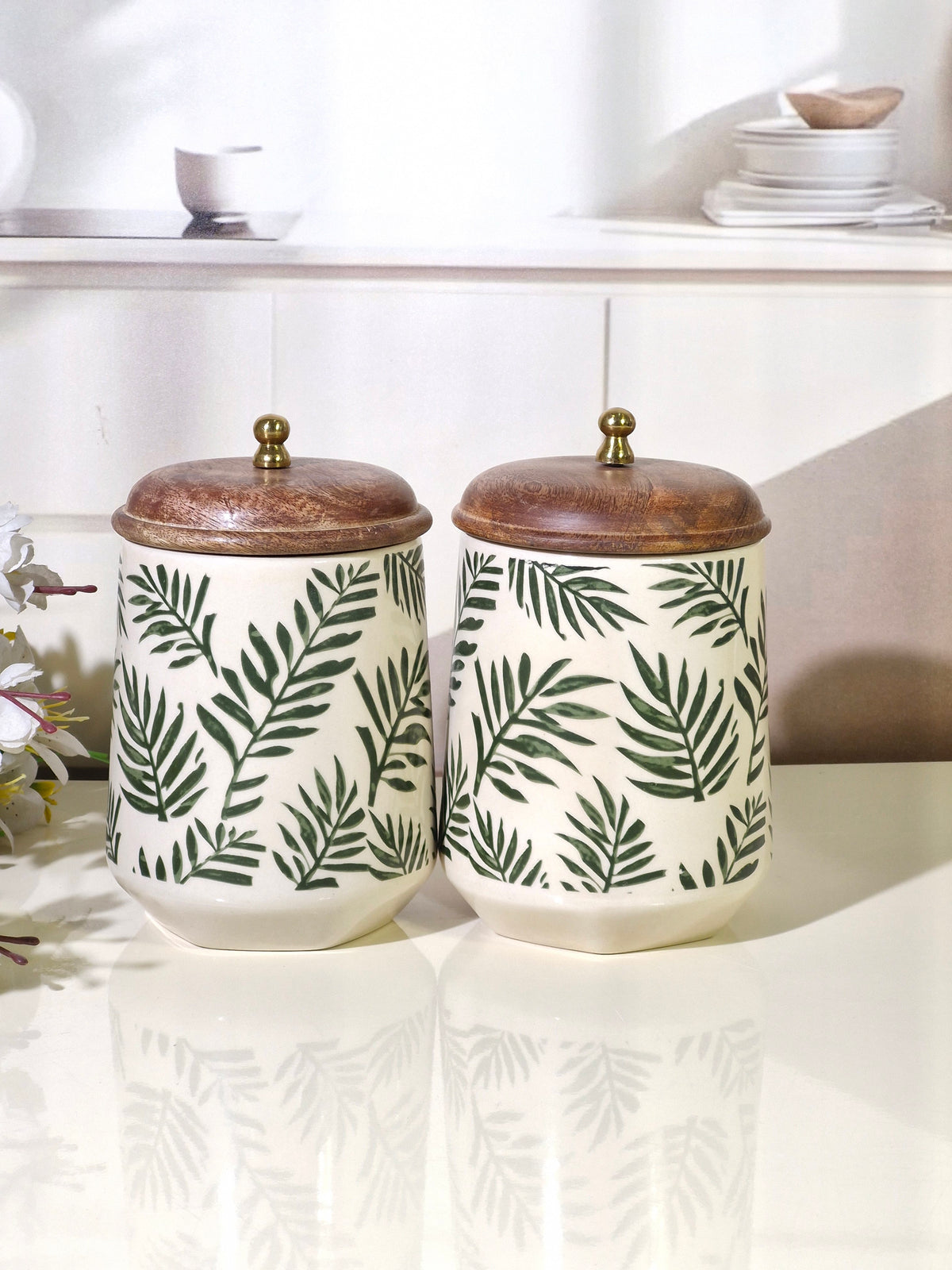 Two ceramic canisters with green palm leaf patterns on a cream background, featuring brown wooden lids with brass knobs, displayed on a glossy white surface.