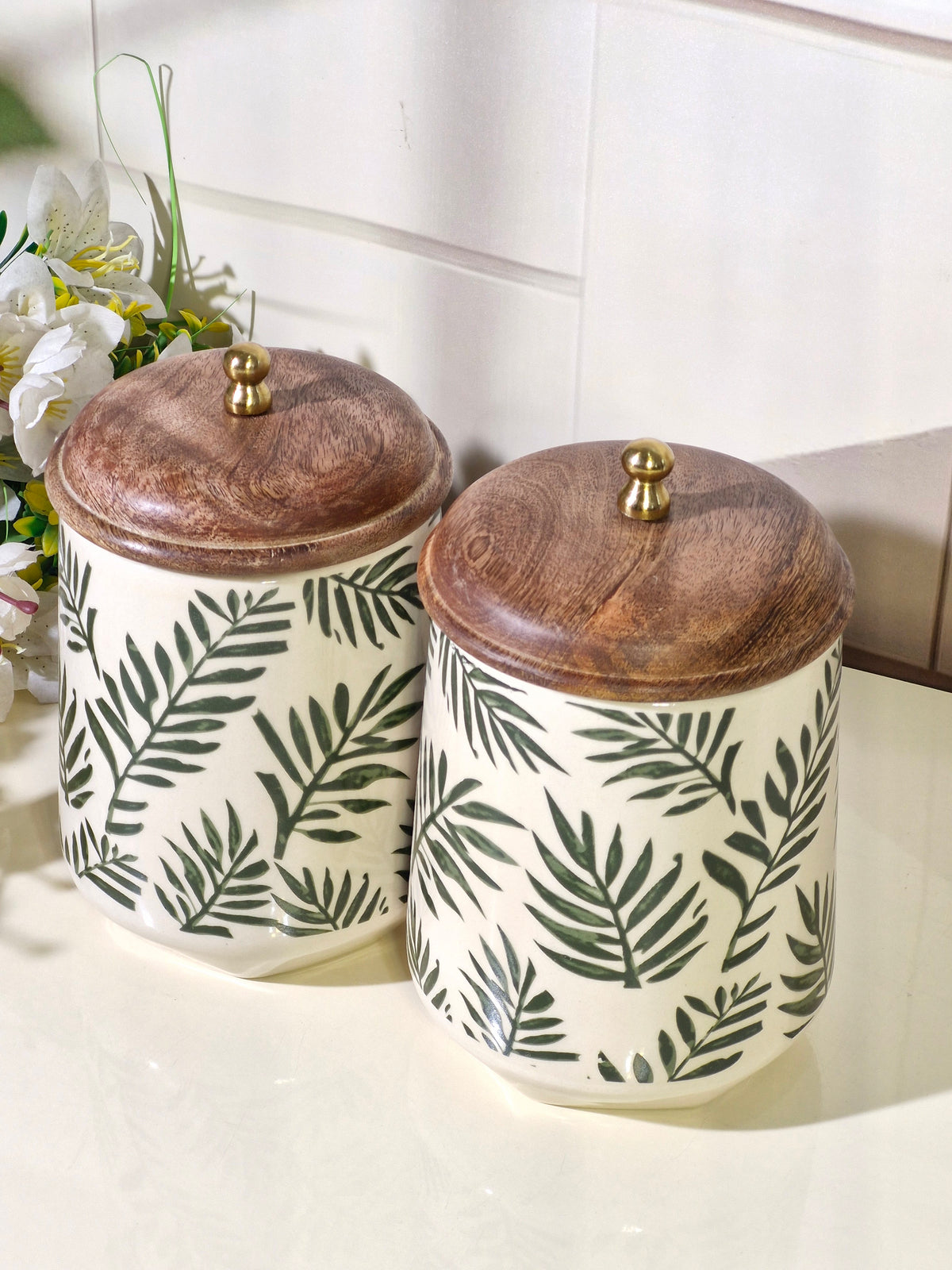 Two cylindrical storage jars with white bodies, dark green palm leaf patterns, and rustic brown wooden lids with small gold knobs, set on a light countertop.