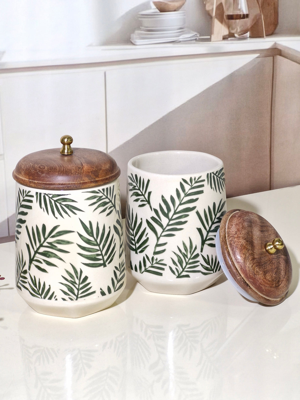 Two ceramic canisters with a green palm leaf pattern on a white background, featuring wooden lids with brass knobs, displayed on a kitchen counter.