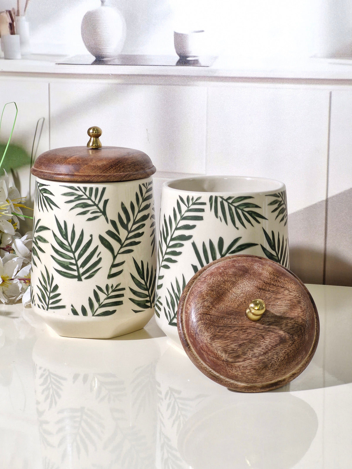 Two ceramic canisters with a green palm leaf pattern on a cream background, featuring dark brown wooden lids with brass knobs, displayed on a glossy white surface.
