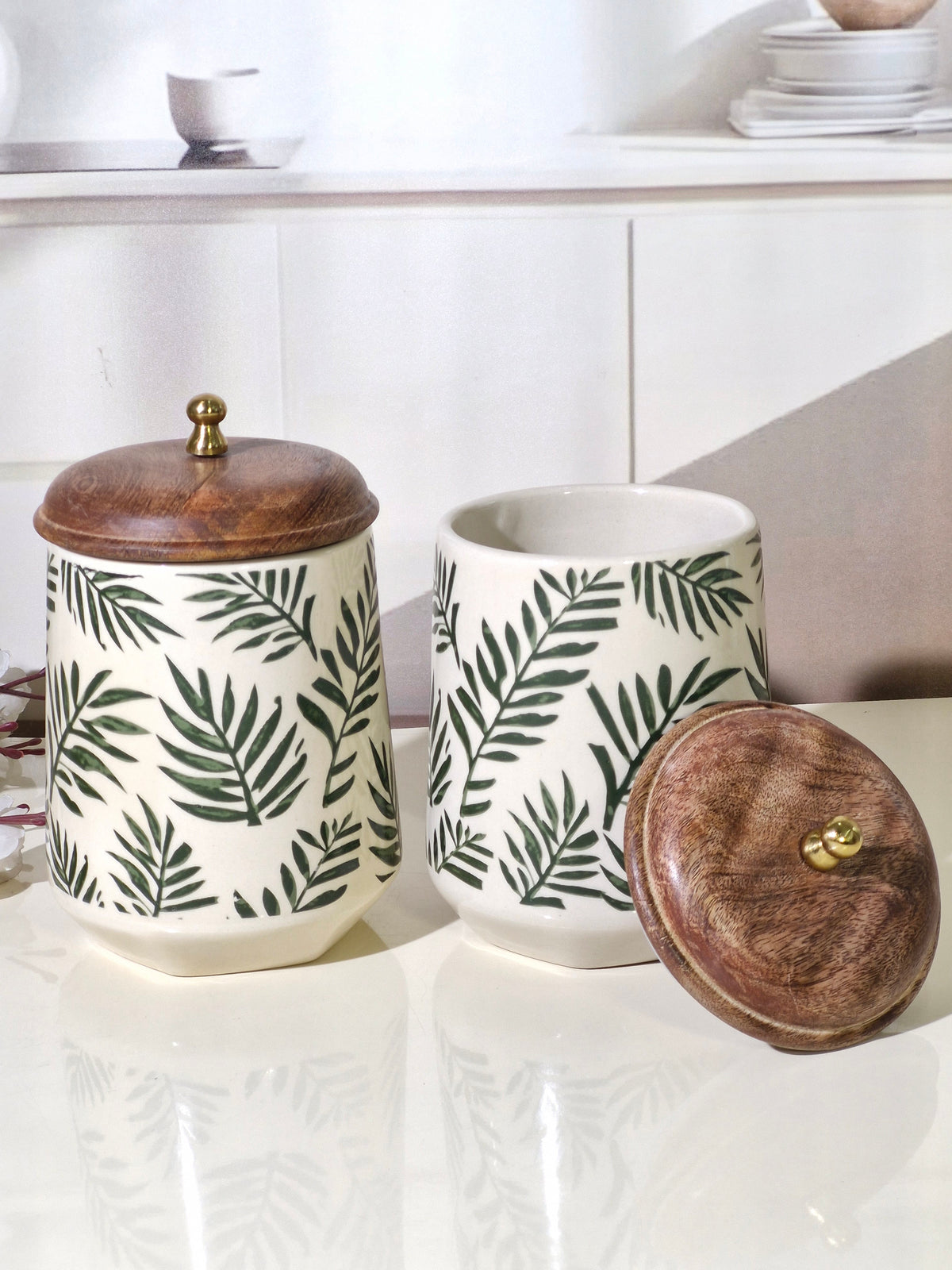 Two ceramic canisters with a green palm leaf pattern on a white background, featuring dark brown wooden lids with brass knobs, displayed on a glossy white countertop.