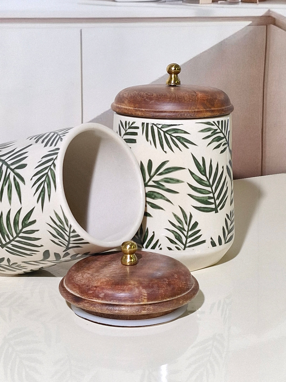 Two ceramic canisters with a green palm leaf pattern on an off-white background, featuring dark brown wooden lids with brass knobs, displayed on a light-colored countertop.