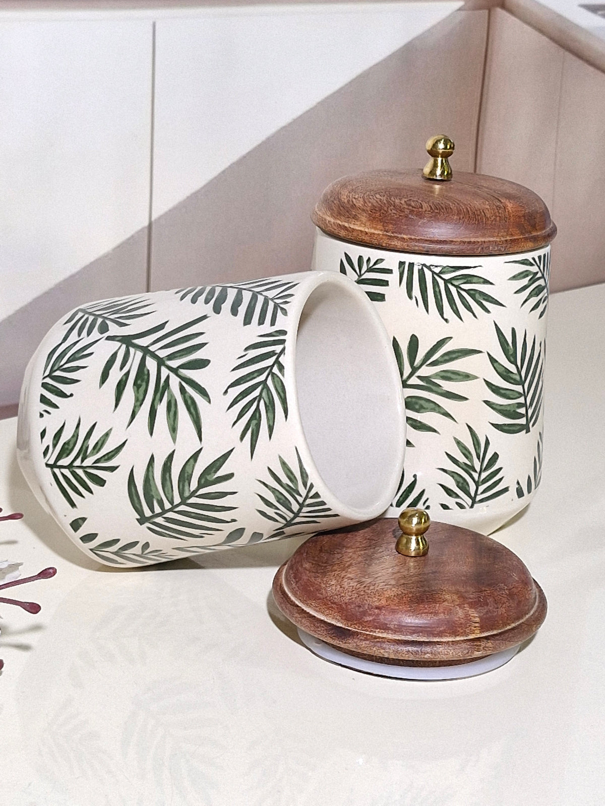 Two ceramic canisters with a green palm leaf pattern on an off-white background, featuring dark brown wooden lids with brass knobs, displayed on a light surface.